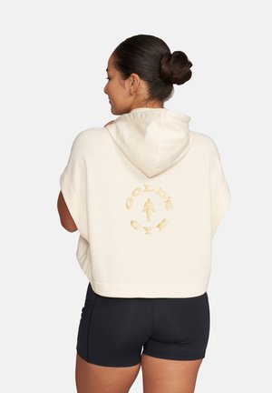 Frau mit Dutt, die eine cremefarbene, ärmellose, kürzere Kapuzenjacke mit dem Gold's Gym-Logo auf dem Rücken und schwarze Sportshorts trägt.