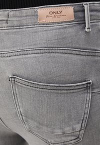 Los jeans de denim gris presentan un acabado texturizado, costuras prominentes y un solo bolsillo trasero. Una etiqueta de cuero dice "ONLY Blue Denim."