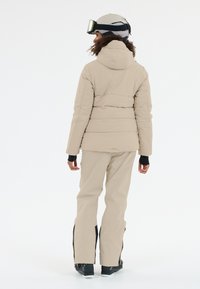 Beige gepolsterte Skijacke mit Kapuze, kombiniert mit beigen Skihosen. Merkmale sind Reißverschlussärmel und ein elegantes Design. Sichtbare schwarze Skischuhe.