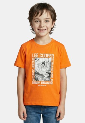 Garçon souriant portant un t-shirt orange avec du texte blanc et un graphique de tigre en noir et blanc, associé à un jean bleu.