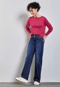 Fuchsia Langarmshirt, kombiniert mit lockeren blauen Denim-Jeans. Die Jeans haben eine hohe Taille, helle Nähte und Taschen auf der Vorderseite. Die Sneaker sind beige.