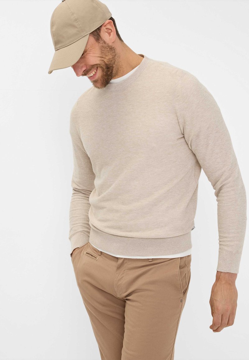 Beige Pullover mit strukturiertem Strick, Rundhalsausschnitt und gerippten Bündchen. Kombiniert mit hellbraunen Hosen und einer passenden Mütze.