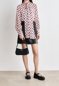 Blouse rose clair à pois noirs, col noué, jupe en simili cuir noir, sac à main noir avec le logo "HUGO", chaussures noires chunky.