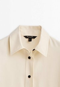 Lys beige skjorte med knapper og spidse kraver, sorte knapper og synlige sømdetaljer. Materialet ser glat og blødt ud.