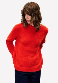 Roter, strukturierten Pullover mit rundem Halsauschnitt und Raglanärmeln. Getragen mit dunkelblauen Jeans. Der Stoff wirkt weich und gemütlich.