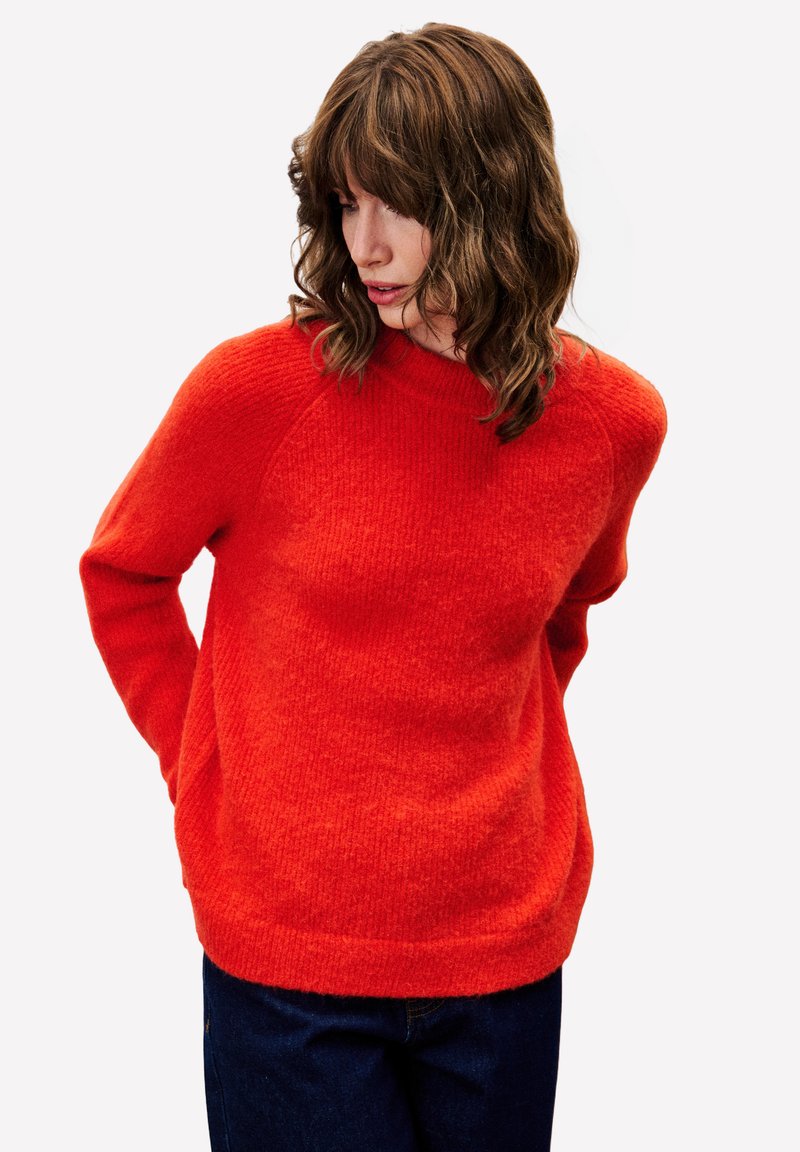 Roter, strukturierten Pullover mit rundem Halsauschnitt und Raglanärmeln. Getragen mit dunkelblauen Jeans. Der Stoff wirkt weich und gemütlich.