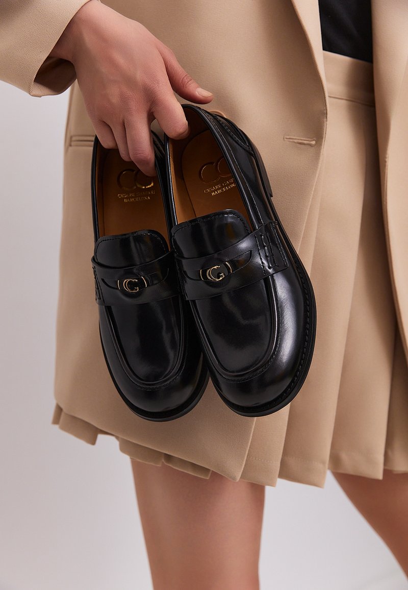 Black leather loafers with a shiny finish, featuring a decorative strap with a gold accent, held in a hand against a neutral background.