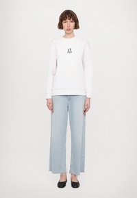 Biela mikina s centrálnym čiernym logom, kombinovaná s svetlomodrými širokými džínsami s rozstrapkanými lemami. Čierne balerínky dopĺňajú outfit.