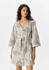 Donna che indossa un vestito beige chiaro con cintura, decorato con motivi floreali e uccelli, maniche a tre quarti e bottoni sul davanti, mentre guarda verso destra.