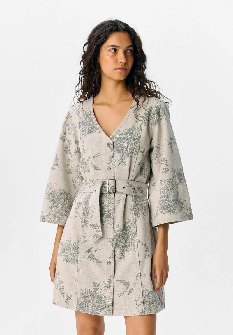 Donna che indossa un vestito beige chiaro con cintura, decorato con motivi floreali e uccelli, maniche a tre quarti e bottoni sul davanti, mentre guarda verso destra.