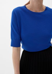 Blå kortärmad stickad topp med ribbade manschetter, inskjuten i en högmidjad svart plisserad kjol på en person.