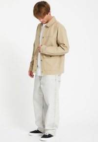 Chaqueta de lienzo beige con cierre de botones y dos bolsillos en el pecho, llevada sobre una camisa blanca y combinada con jeans gris claro y zapatillas negras.