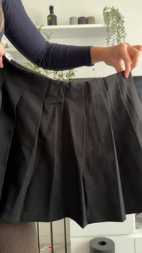 Jupe noire plissée avec une texture lisse et une ceinture. Présente plusieurs plis verticaux qui augmentent le volume à l'ourlet.