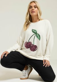 Blondynka klęczy, ubrana w biały sweter z cekinowym wzorem wiśni, czarne spodnie i białe trampki na jednolitym tle.