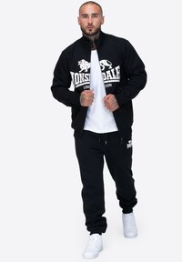 Czarna kurtka zapinana na zamek z logo "Lonsdale London", biała koszulka, czarne dresy i białe tenisówki. Gładki materiał, sportowy fason, casualowy styl.