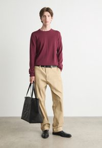 Maglione a maniche lunghe color marrone, pantaloni khaki e scarpe nere. Il modello tiene in mano una borsa a spalla nera con la scritta "HUGO" impressa sul lato.