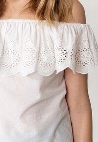 Top bianco scollato con bordo in pizzo ad occhio di pernice ricamato e ondulato. Texture morbida, tessuto leggero, design casual.