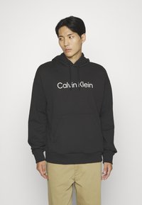 Black hoodie featuring "Calvin Klein" logo in white text, kangaroo pocket, drawstring hood, and ribbed cuffs and hem. Smooth fabric texture.