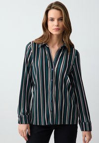 Koszula zapinana na guziki z pionowymi paskami w kolorze teal, czarnym i kremowym. Długie rękawy z guziczkami oraz kołnierz. Gładka struktura materiału.