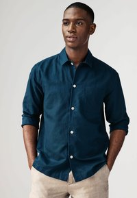 Camisa de botón de color azul marino con un bolsillo en el pecho, mangas enrolladas y botones blancos, hecha de una tela texturizada. Los pantalones son de un beige claro.