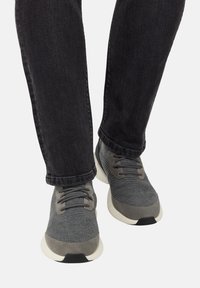 Osoba oblečená v šedých pletených teniskách s bielymi podrážkami a čiernymi šnúrkami, spárovaná s čiernymi džínsami na bielom pozadí.