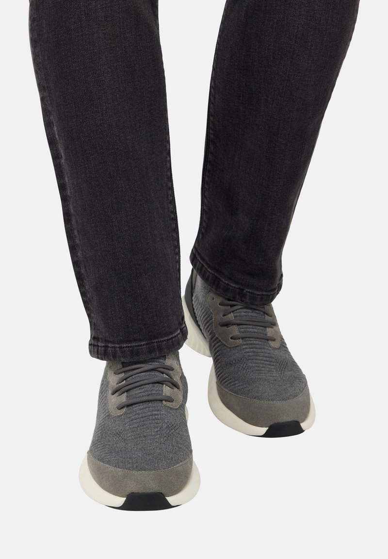 Osoba oblečená v šedých pletených teniskách s bielymi podrážkami a čiernymi šnúrkami, spárovaná s čiernymi džínsami na bielom pozadí.