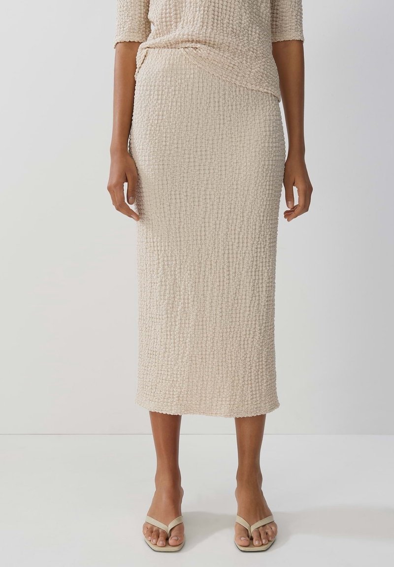 Jupe midi beige texturée portée par une personne debout, associée à un haut assorti et des sandales beiges à bouts ouverts avec de fines lanières.