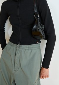 Person som bär en svart ribbad zip-up tröja, gröna byxor med elastisk midja och bär en liten svart läderväska över axeln.