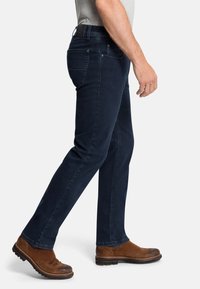 Dunkelblaue Jeans mit slim-fit, aus Denim gefertigt, mit fünf Taschen und dezenten Stickdetails; getragen mit braunen Wildlederschuhen.