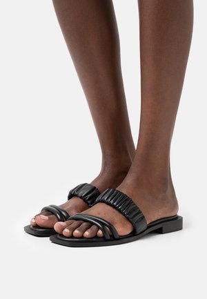 Zwarte leren slippers met een gepolsterde bovenkant met geplooide details en twee in elkaar verweven banden. Platte zool met een vierkante teenvorm.