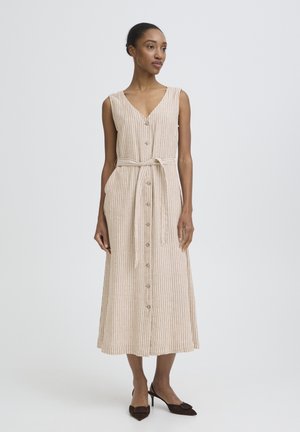 Donna che indossa un abito midi senza maniche beige e bianco a righe, con bottoni sul davanti e cintura annodata in vita, abbinato a tacchi neri.