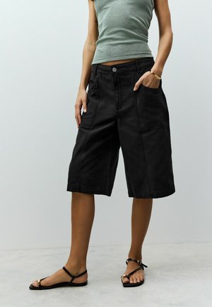 Black denim culottes with a relaxed fit, featuring front pockets and stitched details. Worn with a green ribbed tank and flat sandals.