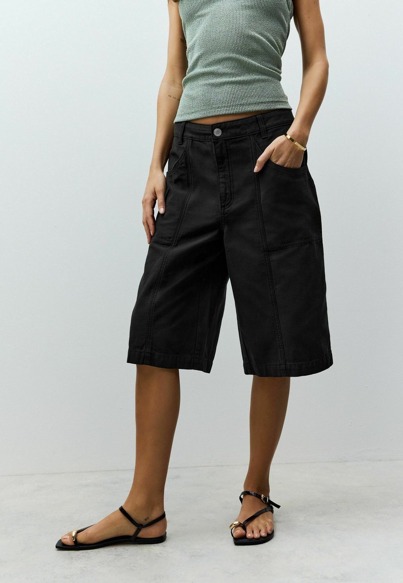 Schwarze Denim-Culottes mit lockerer Passform, ausgestattet mit Fronttaschen und genähten Details. Tragen kombiniert mit einem grünen gerippten Tanktop und flachen Sandalen.
