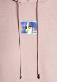Sweat à capuche rose clair avec un patch holographique carré marqué "HUGO" en lettres noires, avec des cordons aux embouts métalliques argentés.
