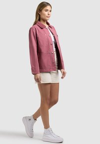 Frau steht, trägt eine staubig rosa geknöpfte Jacke über einem weißen Oberteil, beige Shorts und weiße hohe Sneakers, vor einem schlichten hellen Hintergrund.
