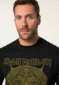 Černé bavlněné tričko s propracovaným zeleným grafickým designem a textem "Iron Maiden" s podrobným centrálním motivem.