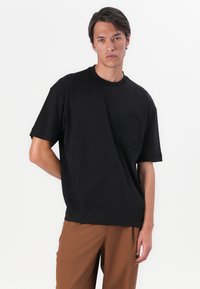 T-shirt en coton noir avec un col rond, des manches courtes et une poche poitrine. Présente un léger design asymétrique.