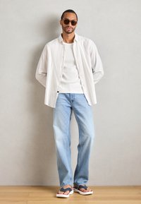 Camicia bianca a bottone sopra una t-shirt bianca, jeans azzurri chiari e infradito nere con accenti bianchi e rossi, su uno sfondo neutro.