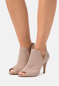 Stivali alla caviglia beige con punta aperta, realizzati in morbido camoscio, caratterizzati da un design curvo, un tacco affusolato e una silhouette minimalista.