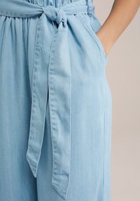 Ljusblå denim jumpsuit med knytbart midjebälte, sidofickor och mjukt, texturerat tyg med avslappnad passform.