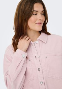 Jonge vrouw met bruin haar die een lichtroze corduroy jasje met knopen draagt, kijkt naar de zijkant terwijl ze de kraag vasthoudt.