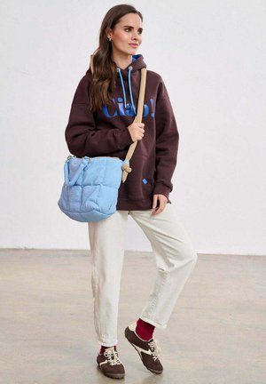 Jasnoniebieska pikowana torba typu bucket z krótkimi uchwytami i długim paskiem. Modelka ubrana w brązową bluzę, białe spodnie i brązowe trampki.