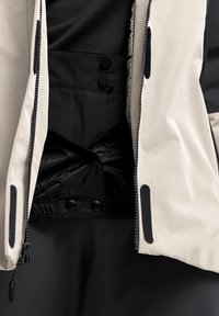 Giacca beige chiaro con zip, interno nero e bottoni in tessuto. Presenta texture a contrasto e un design strutturato. Accenti neri visibili.