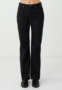 Pantaloni a zampa in denim nero con vita a mezza altezza, due tasche frontali e dettagli in metallo lucido. Il tessuto è liscio con una leggera elasticità.