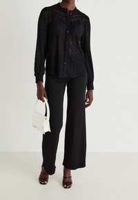 Blouse en dentelle noire à manches longues avec fermeture boutonnée. Associée à un pantalon noir ample et un sac à main blanc. Sandales noires à lanières.