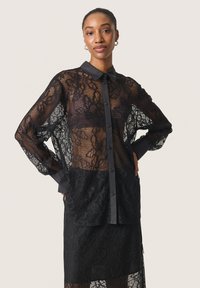 Top in pizzo nero con design trasparente, chiusura a bottoni sul davanti e colletto e polsini in tinta unita a contrasto. Presenta motivi floreali e una vestibilità comoda.