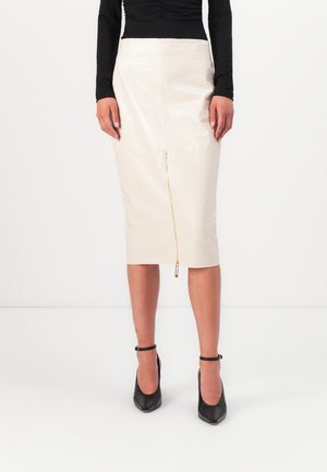 Femme portant une jupe midi blanche effet crocodile avec une fermeture éclair à l'avant, un top noir à manches longues et des talons noirs à brides autour de la cheville.