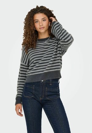 Young woman with curly hair wearing gray and black striped sweater and dark blue jeans, posing with one hand touching her hair.