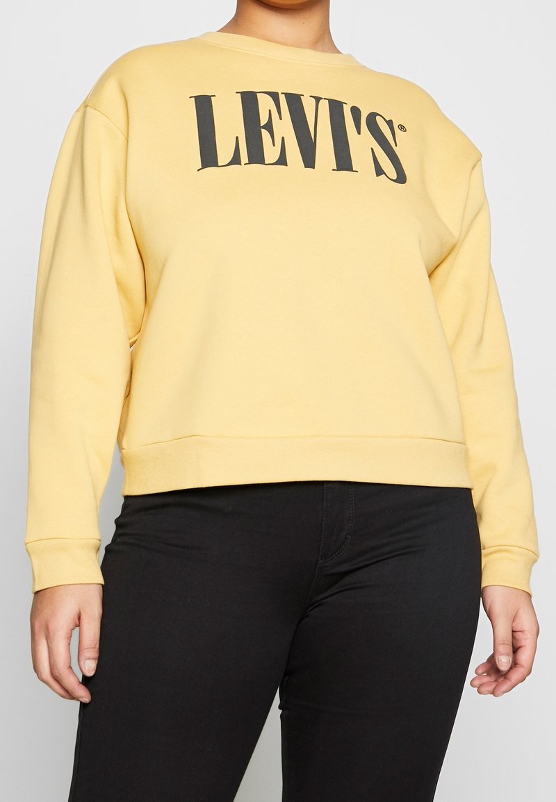 Sudadera amarilla de Levi's con un gran logo negro, cuello redondo, mangas largas y dobladillo acanalado, combinada con pantalones negros.