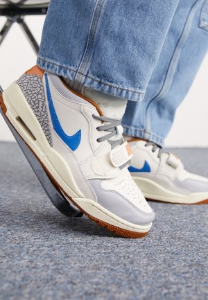Person iført hvide og grå sneakers med blå swoosh-logoer og brune accenter, løfter den ene fod på et gråt tæppe.